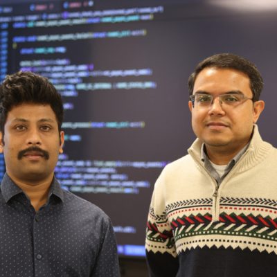 Two men pose in front of a large screen displaying lines of computer code.