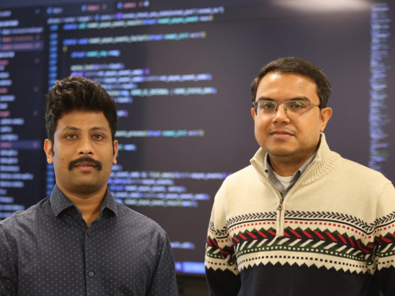 Two men pose in front of a large screen displaying lines of computer code.