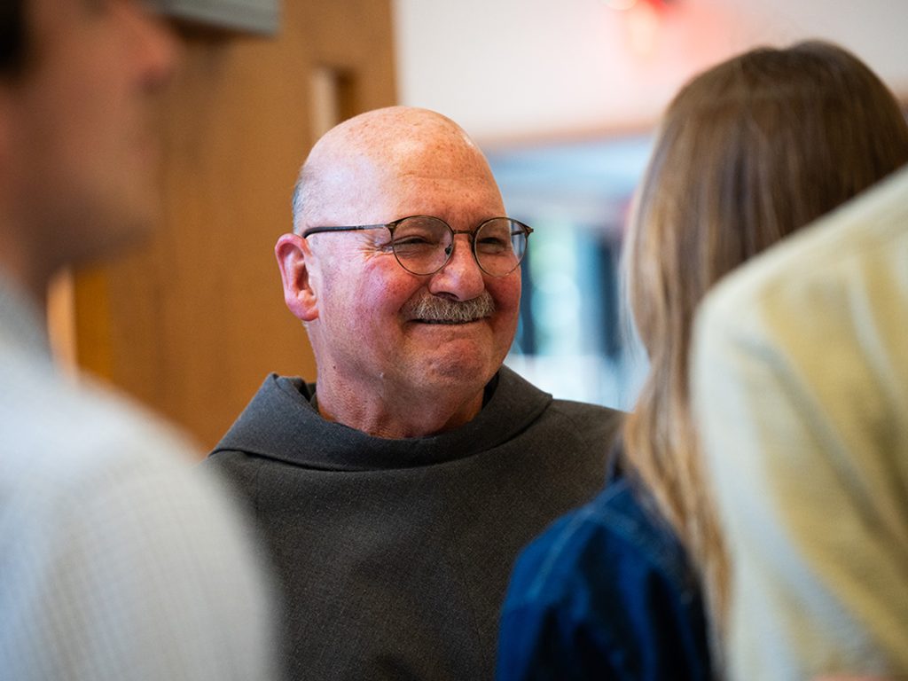 A smiling man wearing glasses and a  brown Franciscan habit converses warmly with people around him at an indoor gathering.