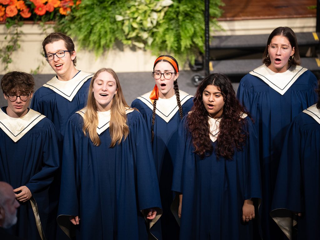 A group of student choir members singing in navy blue robes with cream accents, with floral arrangements visible in the background.