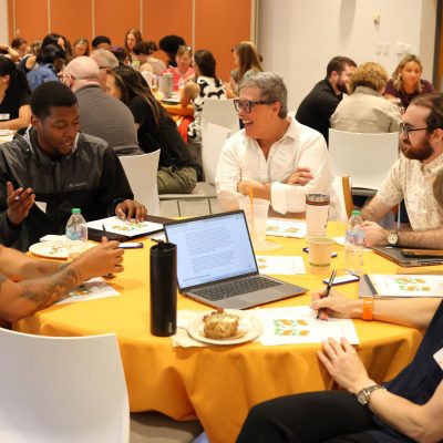 People seated at round tables in a conference room, engaged in small-group discussion with laptops, notebooks and coffee