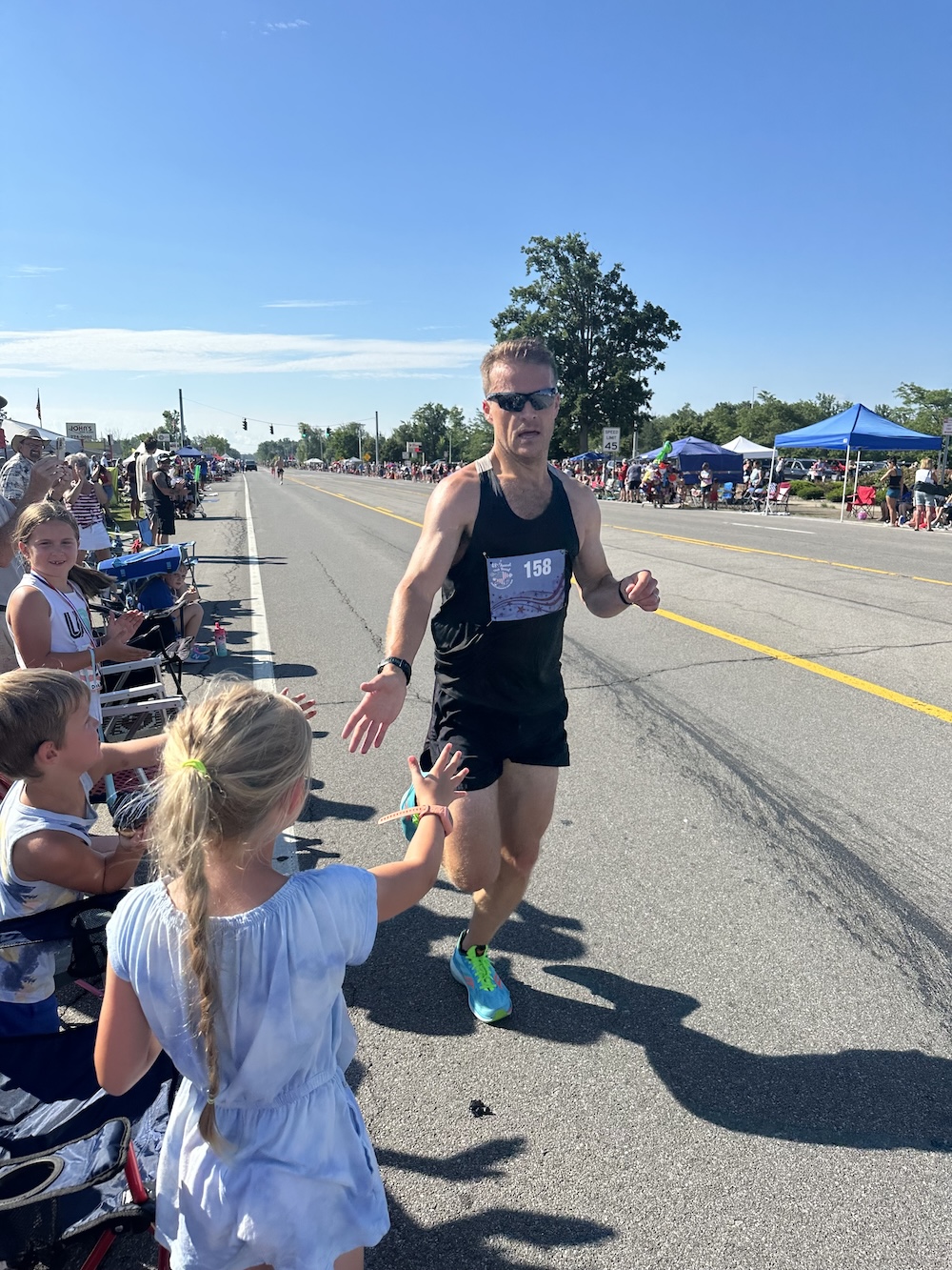 Runner in a black race outfit reaches out to high-five a child along a parade-lined street during a road race.