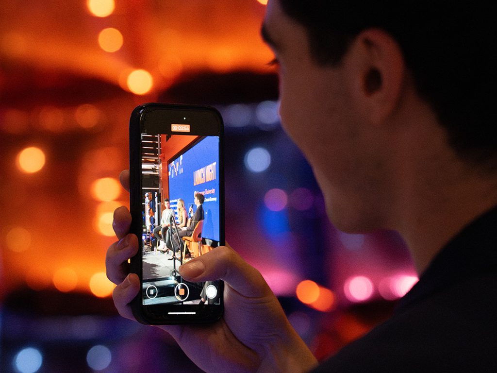 A person films a panel discussion on their smartphone at a Syracuse University Launch Night event, with bokeh lights glowing in the background.