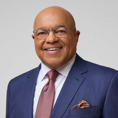 Portrait of Mike Tirico wearing a navy suit, white dress shirt, patterned tie, and glasses against a light gray background.