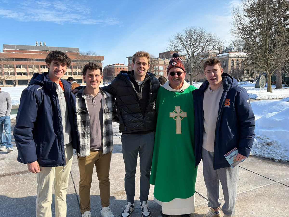 A priest in green vestments and a winter hat poses outdoors with four young men on a sunny, snowy college campus.