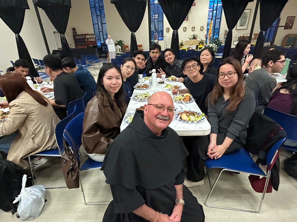A Franciscan priest smiles at the camera while seated at a long dinner table with a diverse group of young people enjoying a communal meal in a church hall.