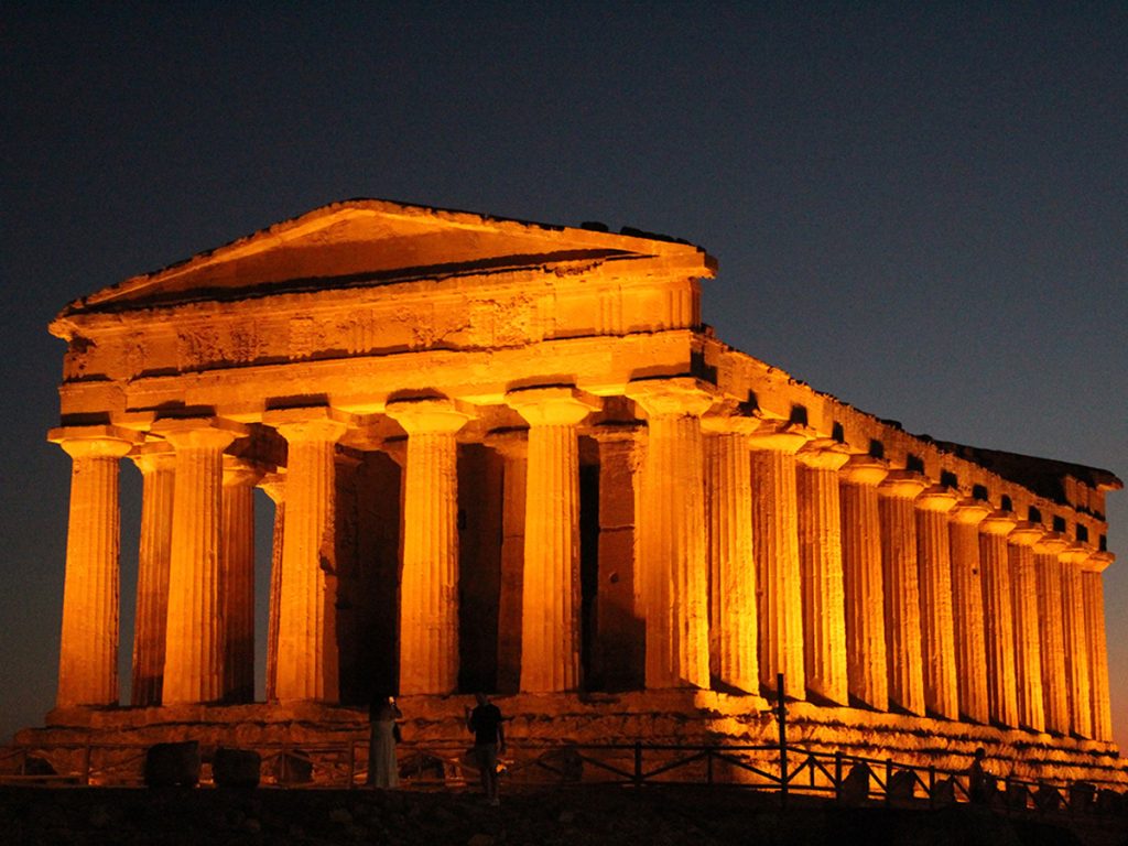 The Temple of Concordia at the Valley of the Temples in Agrigento, Sicily, dramatically illuminated in warm orange light against a dark night sky, with two small figures visible at its base