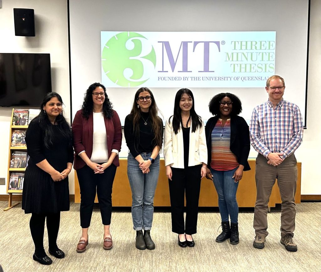 Six people pose together in front of a Three Minute Thesis projection screen in a university library room, including three contest judges and three contestants chosen as winners.