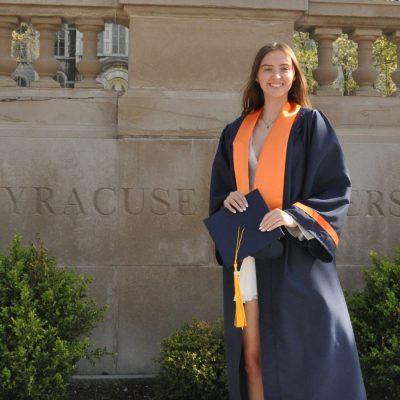A smiling young woman in a navy graduation gown with an orange stole holds her mortarboard in front of a stone wall engraved with "Syracuse University."