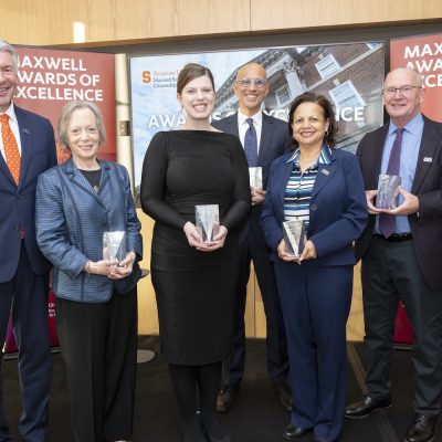 Six people pose holding glass awards in front of “Maxwell Awards of Excellence” signage at a formal ceremony.
