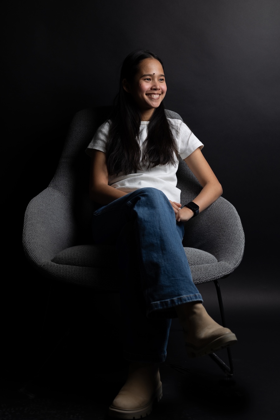 Person seated in a chair against a dark backdrop, wearing a white shirt and jeans.