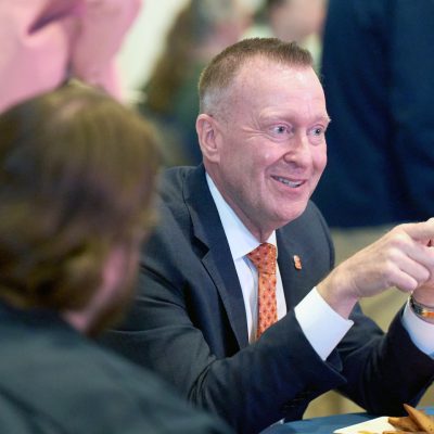 Person in a suit and orange tie gesturing while seated at a table, speaking with others in a casual indoor setting.