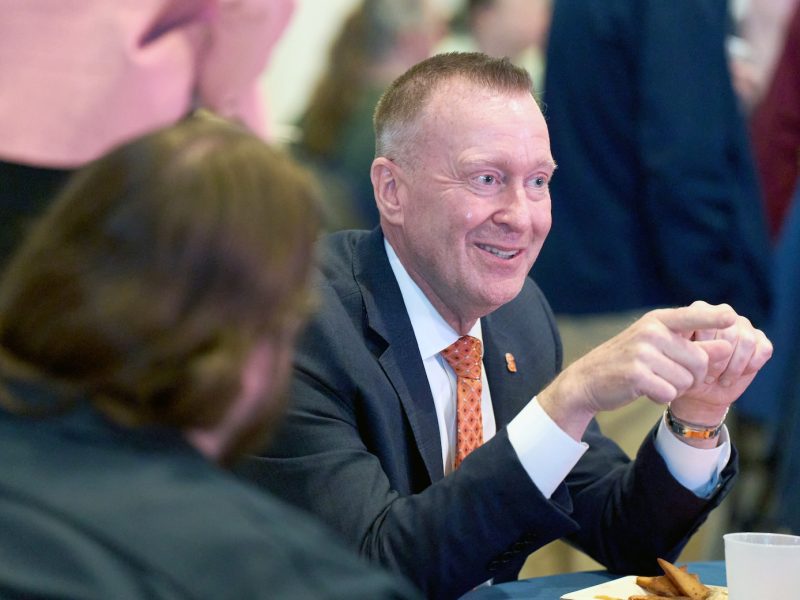 Person in a suit and orange tie gesturing while seated at a table, speaking with others in a casual indoor setting.