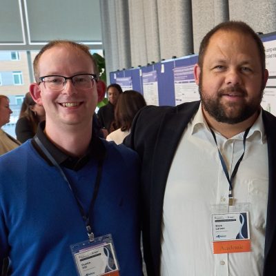 Two conference attendees wearing badges stand together in front of research posters, with other participants and poster boards visible in the background.