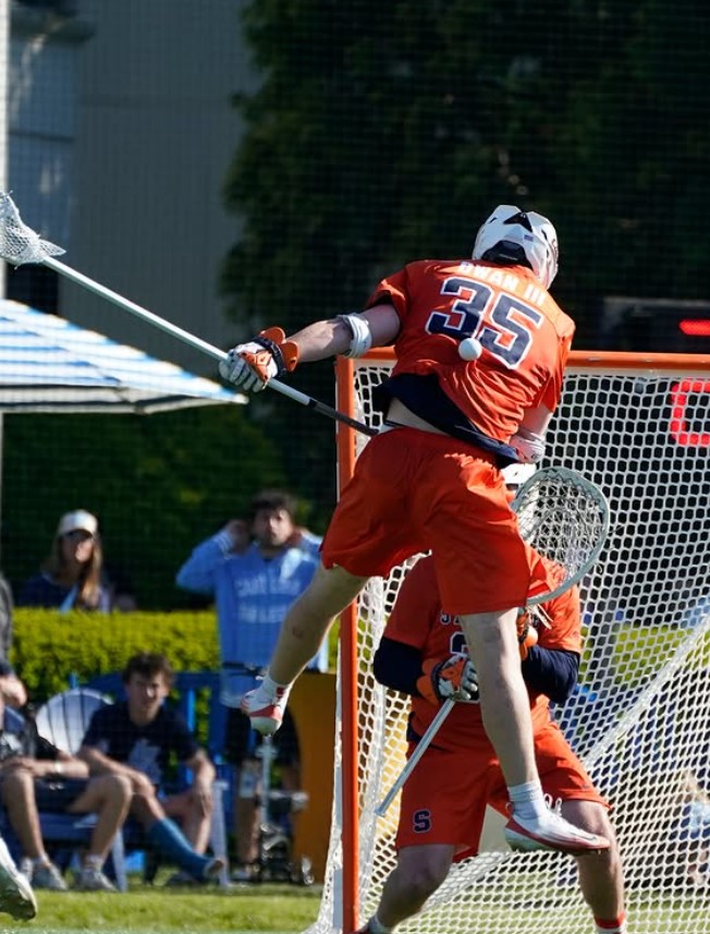 A men's lacrosse player blocks a shot while leaping in the air.