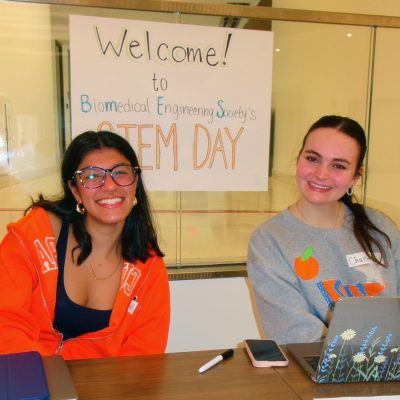 Two smiling students sit at a welcome table in front of a Biomedical Engineering Society STEM Day sign.