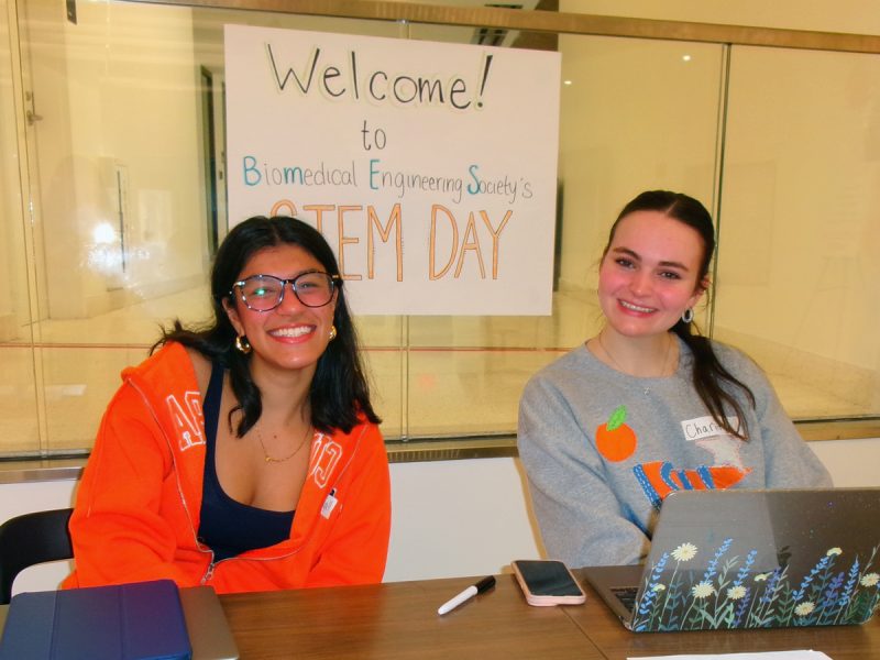 Two smiling students sit at a welcome table in front of a Biomedical Engineering Society STEM Day sign.