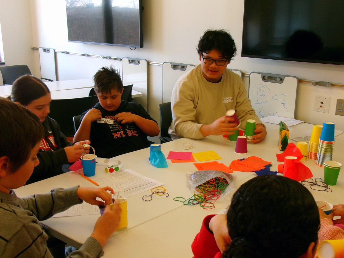 A college student leads young children in a cup-stacking engineering activity using rubber bands and paper.