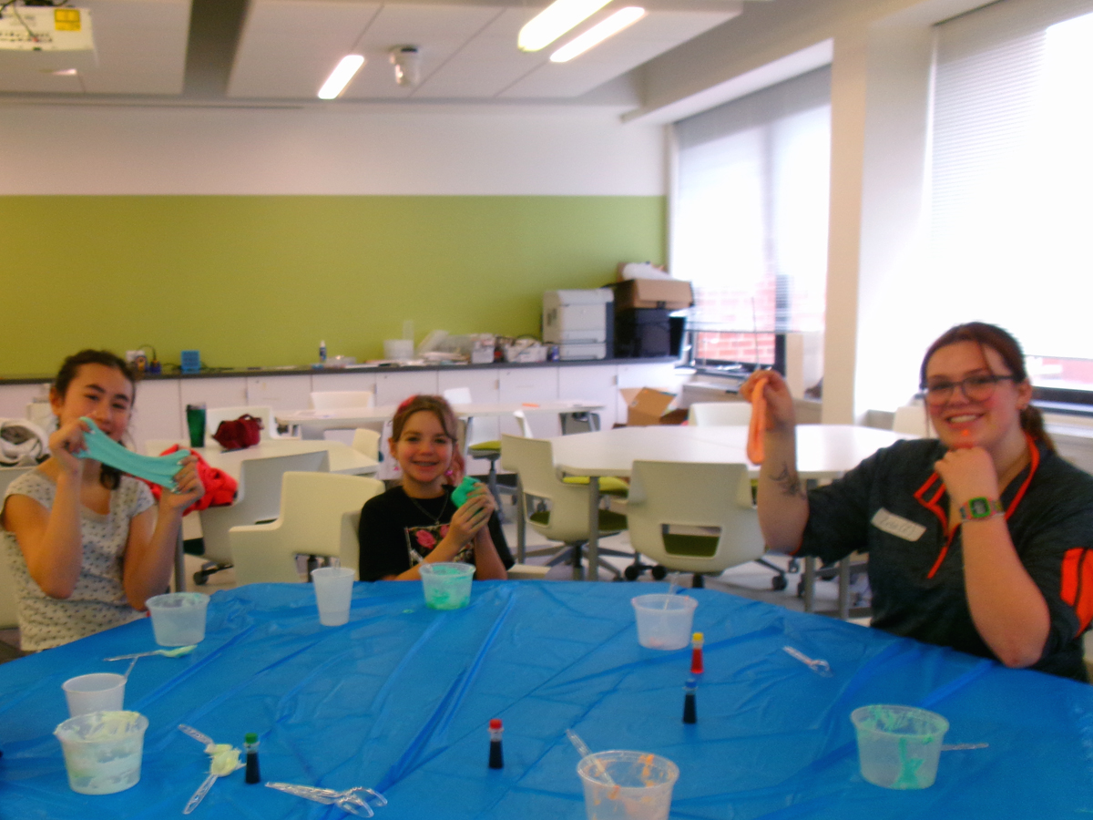 Two children and a college student hold up colorful homemade slime at a table during a STEM activity.