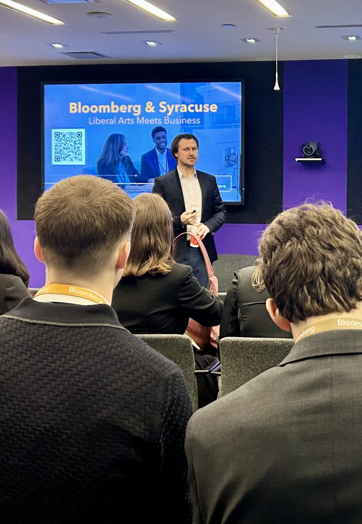 A speaker stands at the front of a conference room before an audience of students wearing Bloomberg lanyards, with a large screen behind him displaying the text 'Bloomberg & Syracuse: Liberal Arts Meets Business."