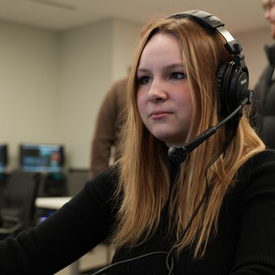 A person wears a headset at an esports broadcast control station, with monitors visible in the background.