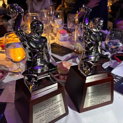 Two silver statuette awards on a dinner table with glasses, candlelight, and plates at an event.