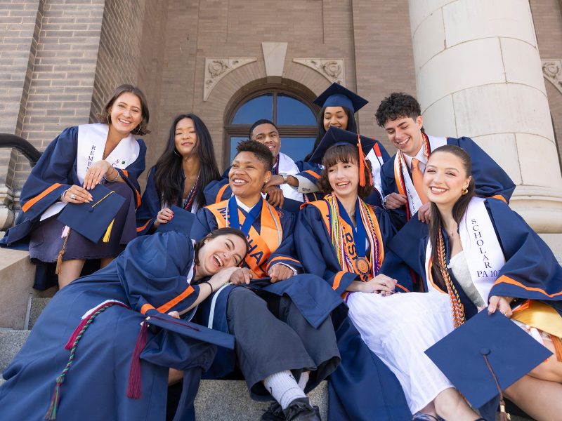 A group of nine Syracuse University graduates in blue and orange caps and gowns laugh and pose together on the stone steps of a campus building