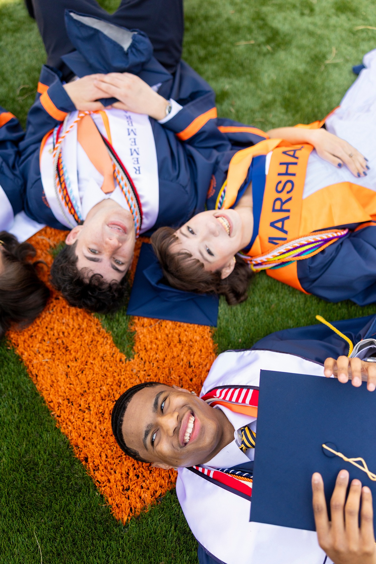 An overhead view of three smiling Syracuse University graduates lying on green turf near an orange block "S," one holding a diploma cover and another wearing a bright orange marshal sash.