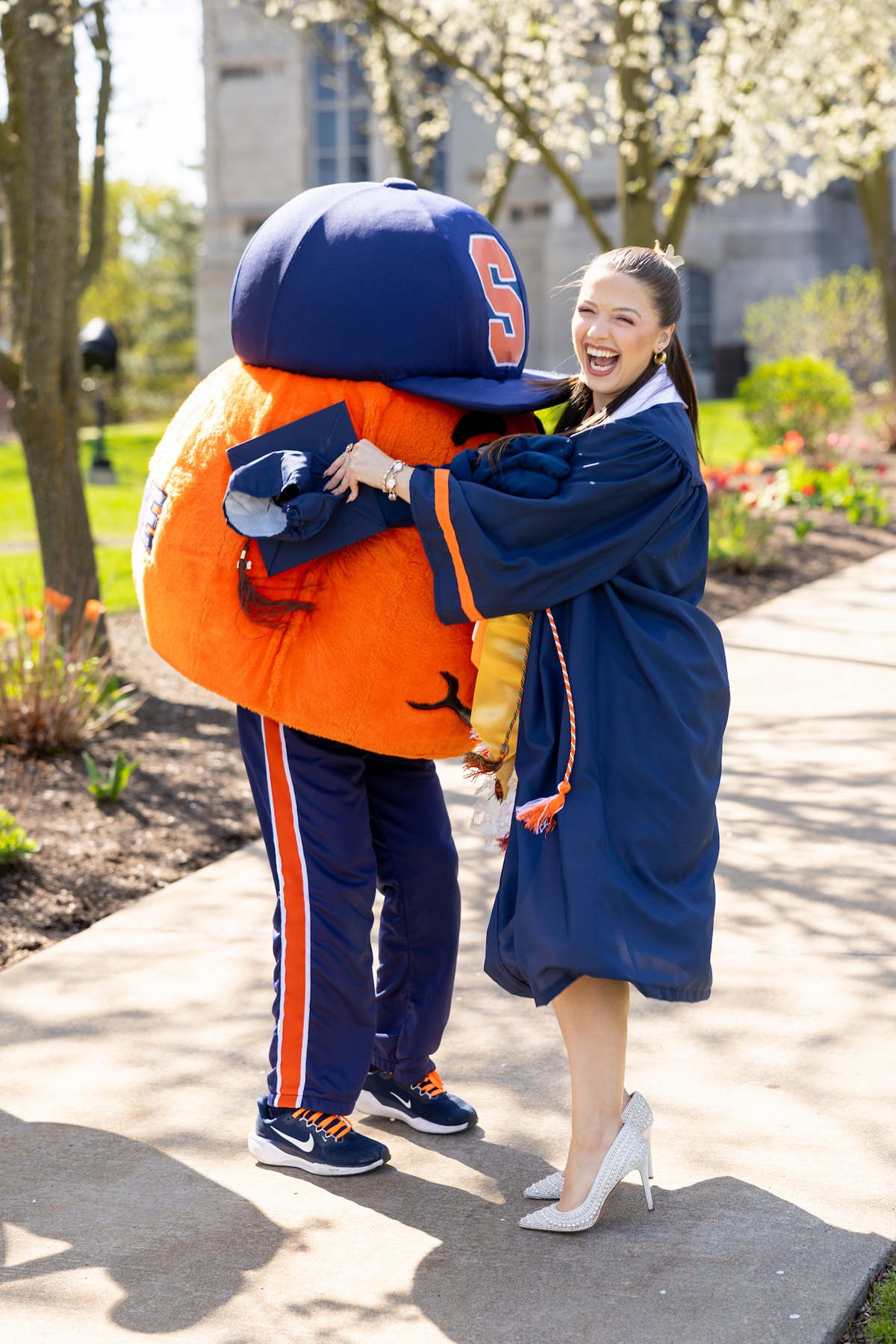 A laughing graduate in a blue cap and gown embraces Otto the Orange on a sunlit campus walkway lined with blooming spring trees.