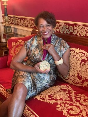 "A woman sits on an ornate red sofa, smiling and proudly displaying a large medallion on a purple ribbon around her neck."
