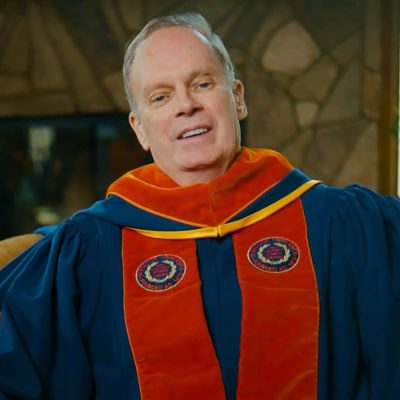 A person wearing Syracuse University doctoral regalia — a blue gown with an orange hood and gold trim, featuring two embroidered University seals — seated indoors with a stone backdrop and greenery visible behind him.