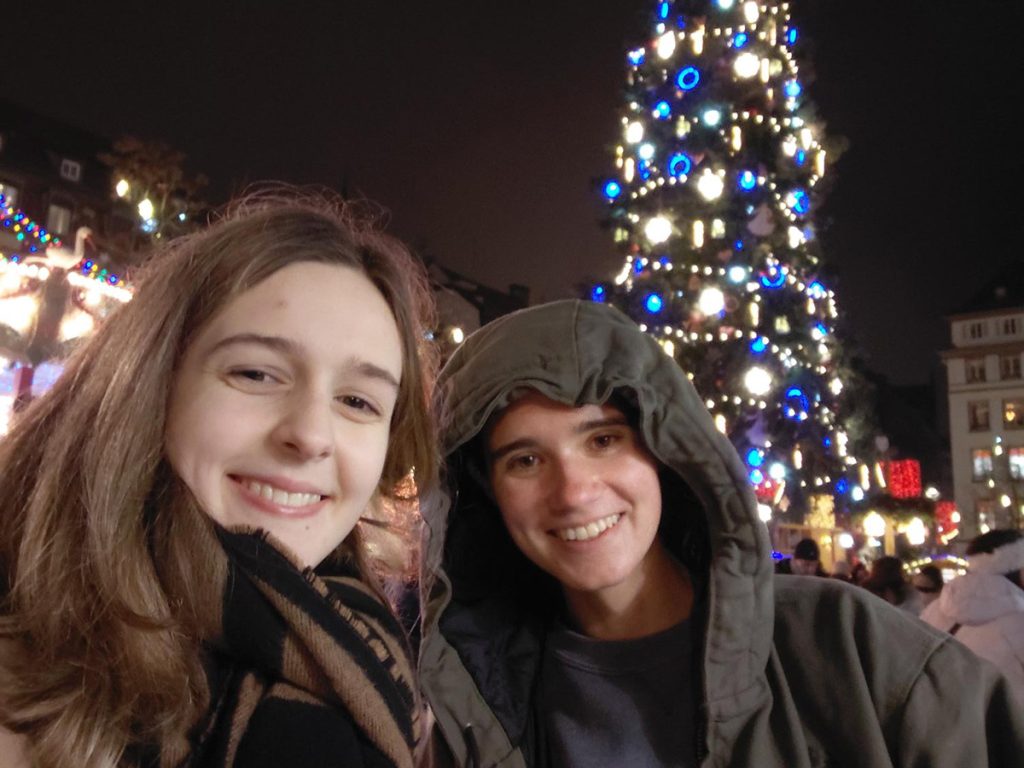 Two young people smile together at a Christmas market at night, with a large decorated tree glowing with white and blue lights behind them."