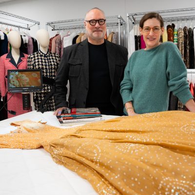 Two people stand behind a white table displaying a golden-yellow beaded or embellished gown spread across its surface. Behind them, metal garment racks hold a variety of stored clothing and textiles