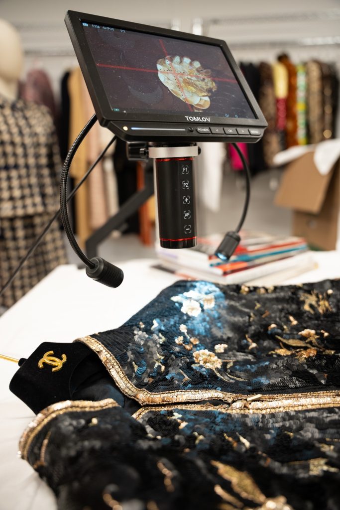 A digital microscope on a flexible stand displays a magnified close-up of fabric details on its monitor screen. Below it, a black and gold embellished garment with sequins and metallic trim lies on a white table.