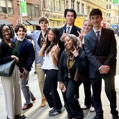 A diverse group of eight professionally dressed young adults pose together on a busy city sidewalk, smiling and making peace signs at the camera. Several are wearing lanyards, suggesting attendance at a professional or academic event.