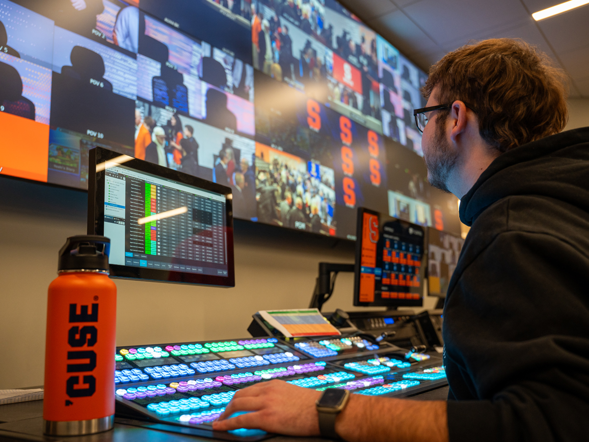 A student sits in front of a broadcast production switcher, with a video wall of esports games playing on the monitors.