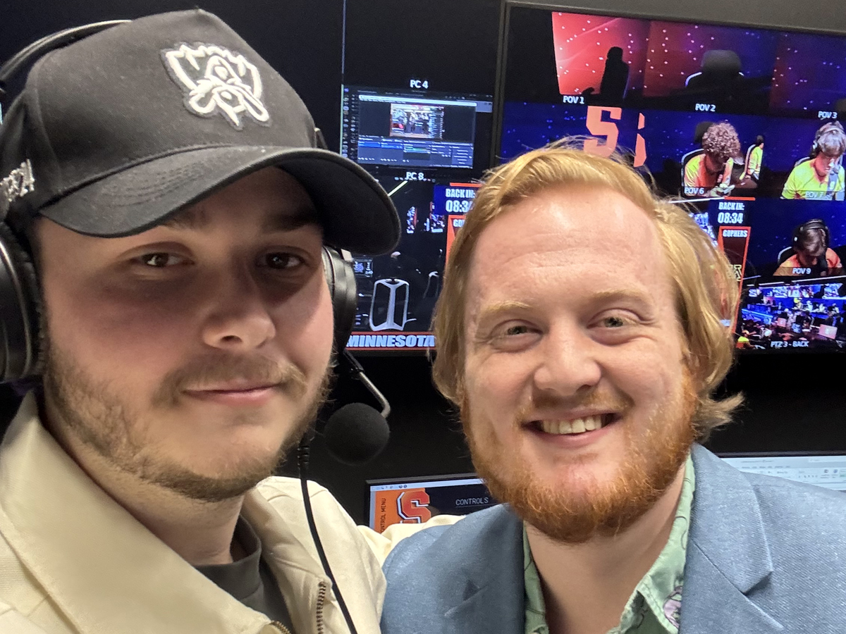 Cole Hilary and Ryan Blankenhorn smile for a selfie at a broadcast production desk with multiple monitors behind them.