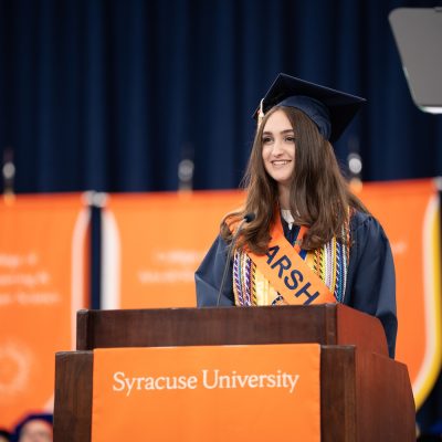 Graduate in cap and gown with an orange sash speaking at a podium labeled “Syracuse University,” with banners in the background.