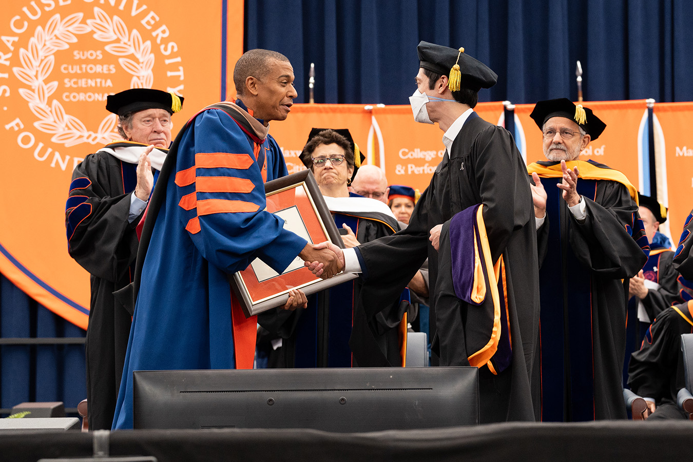 Two people in academic regalia shake hands on stage during a Syracuse University ceremony, with one holding a framed award. Orange banners and the University seal are visible in the background as other faculty and administrators in regalia look on and applaud.