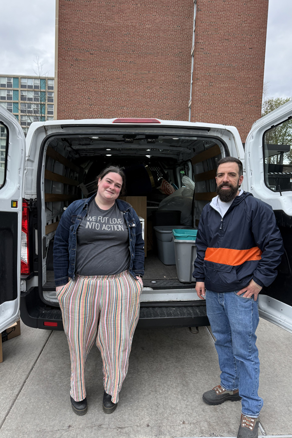 Two volunteers stand at the open rear doors of a van filled with donated items collected during a drive.
