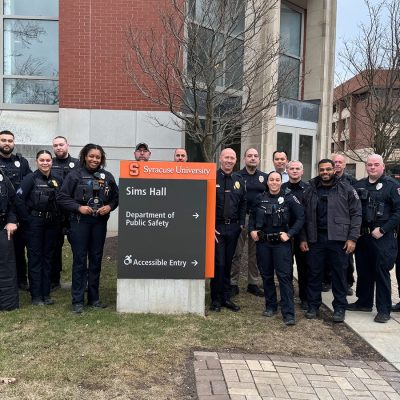 A group of approximately 20 Syracuse University Department of Public Safety officers in uniform pose together outside Sims Hall, standing beside the building's orange-and-gray directional sign