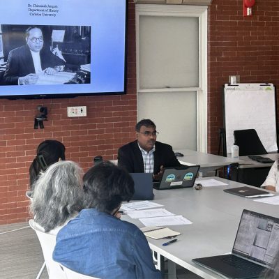 A speaker leads a workshop around a conference table, with a presentation slide titled 'Dalits and Anti-Caste Epistemology' by Dr. Chinnaiah Jangam of Carleton University displayed on the screen behind him