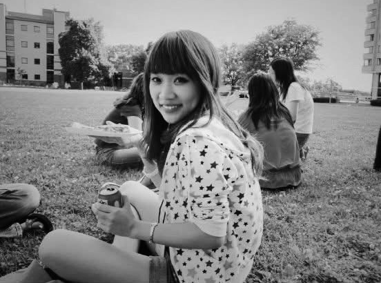 A black-and-white photo of a person sitting on a campus lawn, smiling over their shoulder at the camera while holding a can and a plate of food, with other students and university buildings visible in the background
