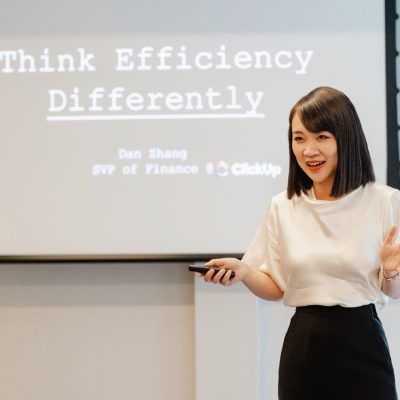 A woman stands in front of a large projection screen, smiling and gesturing mid-sentence while holding a clicker in her right hand. The screen behind her reads "Think Efficiency Differently."
