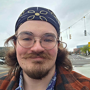 A young man with round glasses, a mustache, and medium-length brown hair wearing a patterned bandana smiles outdoors at a city intersection.