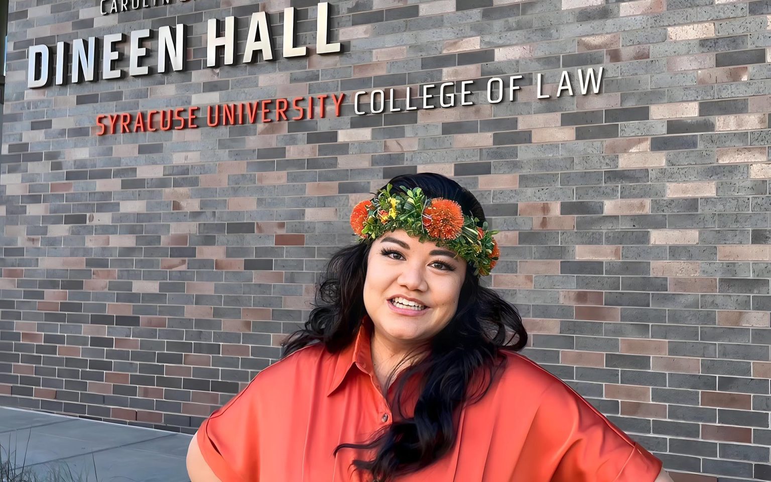 A person wears a floral crown and smiles in front of the Syracuse University College of Law sign.