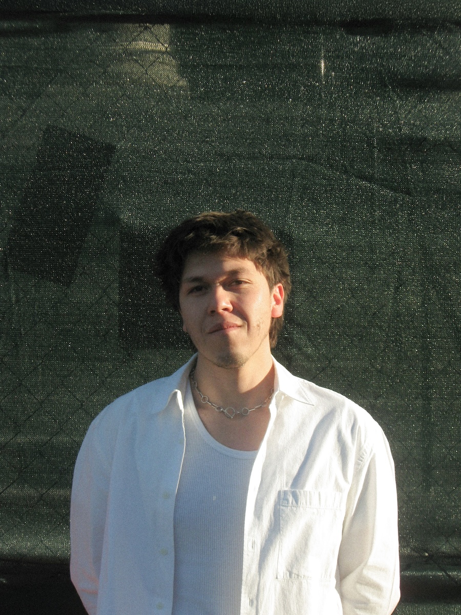 Portrait of a person in a white shirt and necklace standing in sunlight against a dark textured backdrop.
