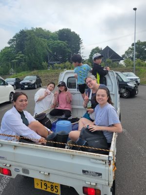 A group of women and children ride together in the bed of a small Suzuki Carry truck in a parking lot, smiling and flashing peace signs, with green trees and a metal structure visible in the background.