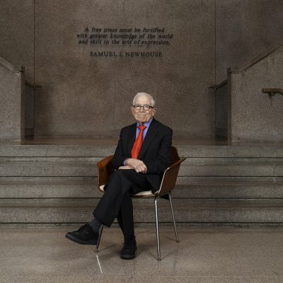 A person in a dark suit and red tie sits in a wooden chair at the base of a stone staircase inside the S.I. Newhouse School of Public Communications. Behind him, a quote from Samuel I. Newhouse is engraved on the wall: "A free press must be fortified with greater knowledge of the world and skill in the art of expression."