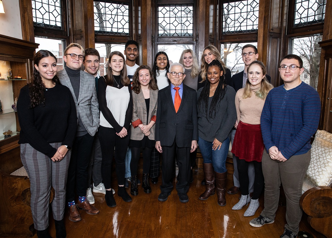 A group of approximately 13 students poses with a person in a dark suit and orange tie in a wood-paneled room with ornate leaded glass windows.